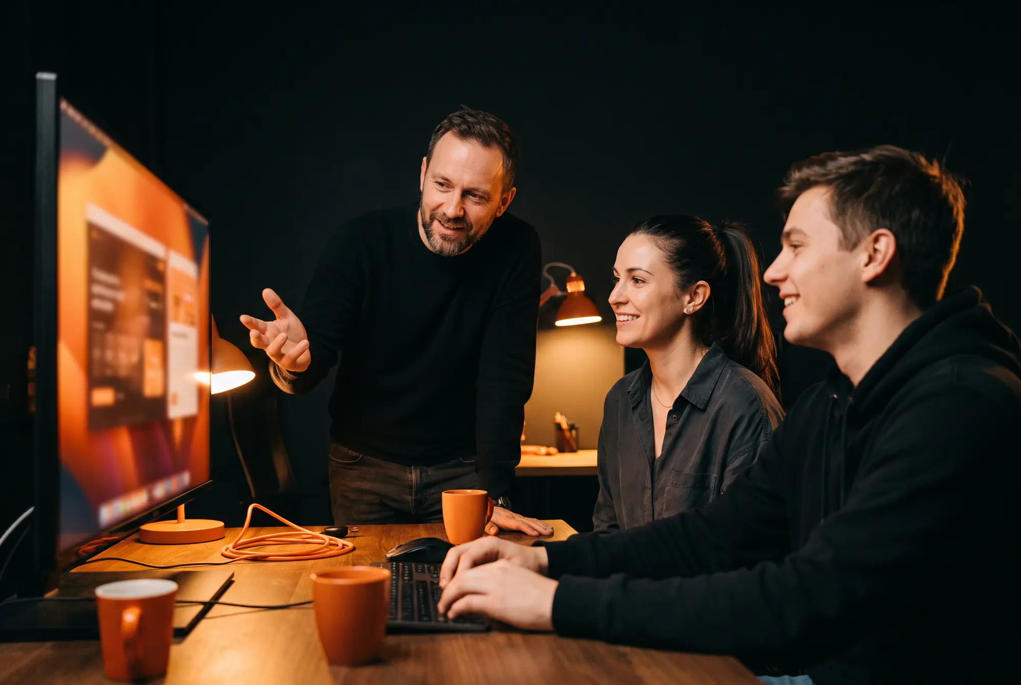 Three teammates smile as they work together at a monitor in a dark studio
