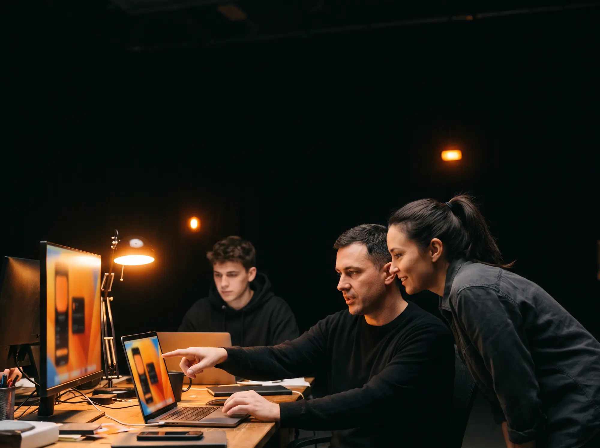 An experienced teammate points at a laptop screen while showing a colleague how it works, a third person works at a monitor behind them